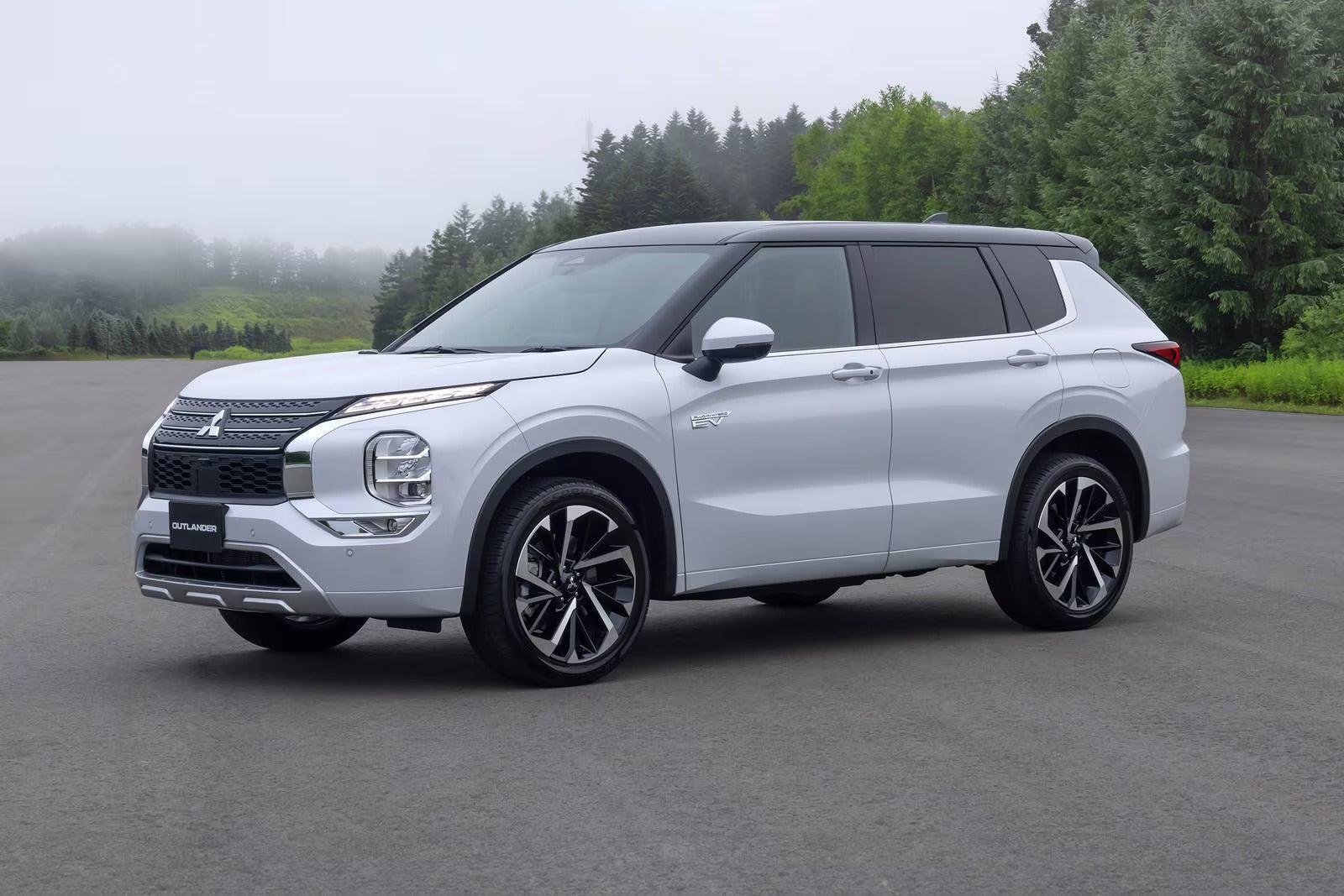 White Mitsubishi Outlander SUV parked on a road with a misty forest background.